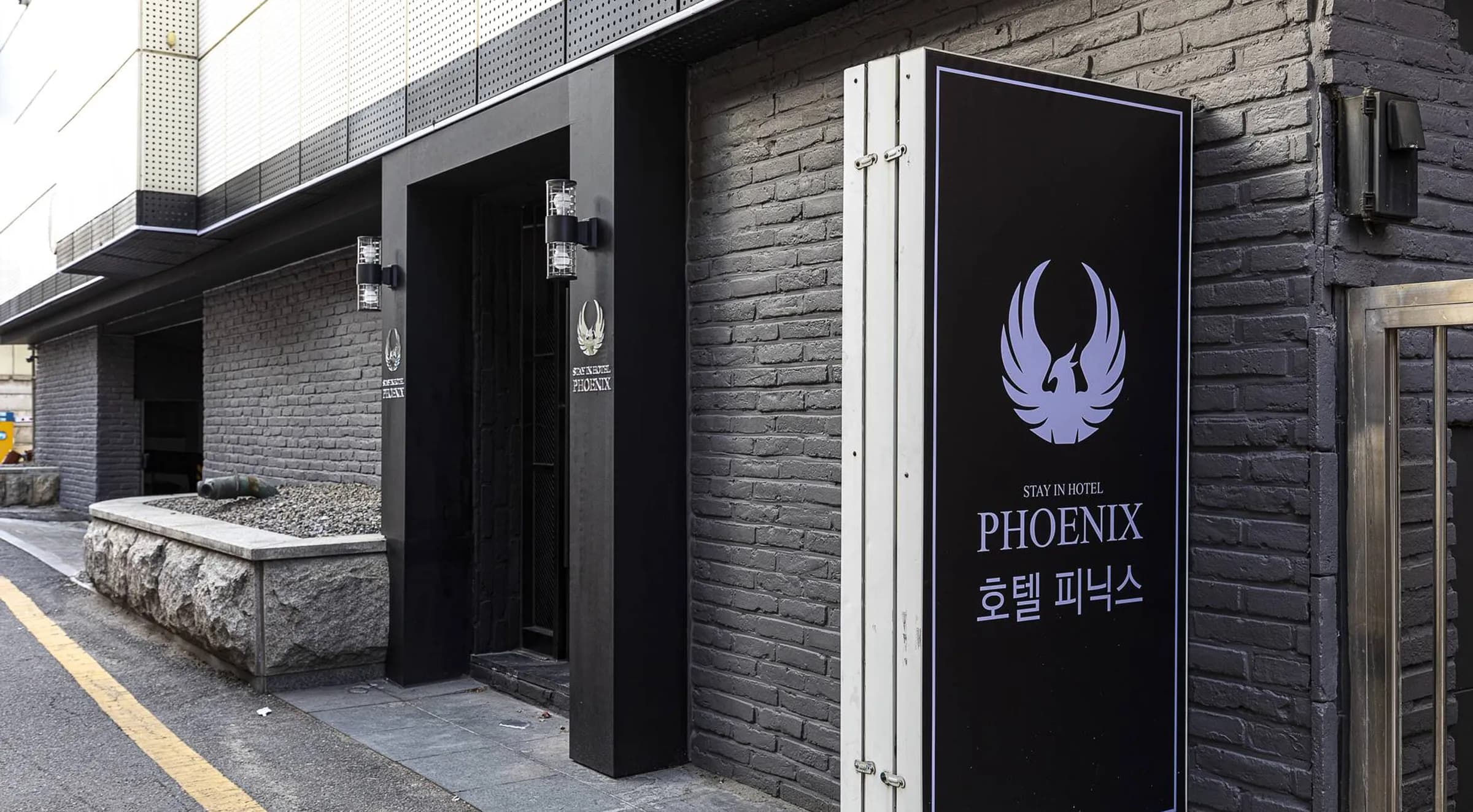 검은색 벽돌 건물 입구에 세워진 불사조 로고와 한국어 이름이 적힌 호텔 피닉스 간판.